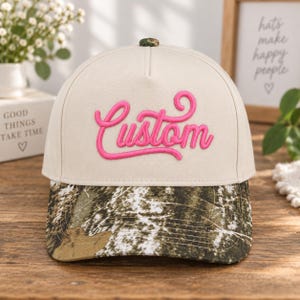 May include: A beige baseball cap featuring a camouflage pattern on the brim and a small button on top. The front panel displays the word Custom embroidered in bright pink cursive script. The cap has a structured crown and a curved visor.