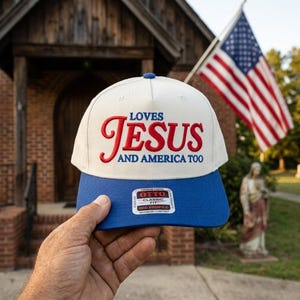 May include: A cream-colored baseball cap with a blue brim featuring the embroidered red and blue text LOVES JESUS AND AMERICA TOO. A rectangular sticker on the brim displays the brand name OTTO, CLASSIC FIT, MID PROFILE, and MAXIMUM COMFORT.