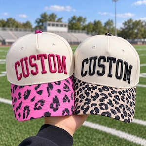 May include: Two baseball caps with off-white crowns and the word CUSTOM embroidered on the front. The left cap features hot pink leopard print on the brim, while the right cap has a tan and black leopard print brim and a matching button.