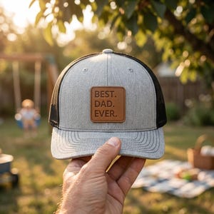 May include: A heather gray baseball cap with a black mesh back and a brown rectangular leather patch on the front. The patch features the embossed text BEST. DAD. EVER. The cap has a structured front panel and a curved brim with stitching.