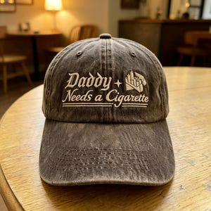 May include: A charcoal gray washed cotton baseball cap featuring the embroidered white text Daddy + Needs a Cigarette with a small graphic of a cigarette pack. The cap has a curved brim, a six-panel construction, and visible stitching details.