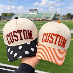 May include: Two baseball caps with cream-colored crowns and the word CUSTOM embroidered on the front. The left cap features a black brim with white polka dots, while the right cap has an orange and white striped brim with an orange top button.
