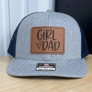May include: A gray and navy blue trucker hat with a leather patch that reads "Girl Dad" in brown lettering. The hat has a curved brim and a mesh back. The Richardson brand label is visible.