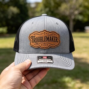 May include: A heather gray and black trucker hat featuring a tan leather-like patch on the front with the word TROUBLEMAKER in a decorative font. The brim has a black sticker with the text RICHARDSON 112 X-LARGE and other small details.