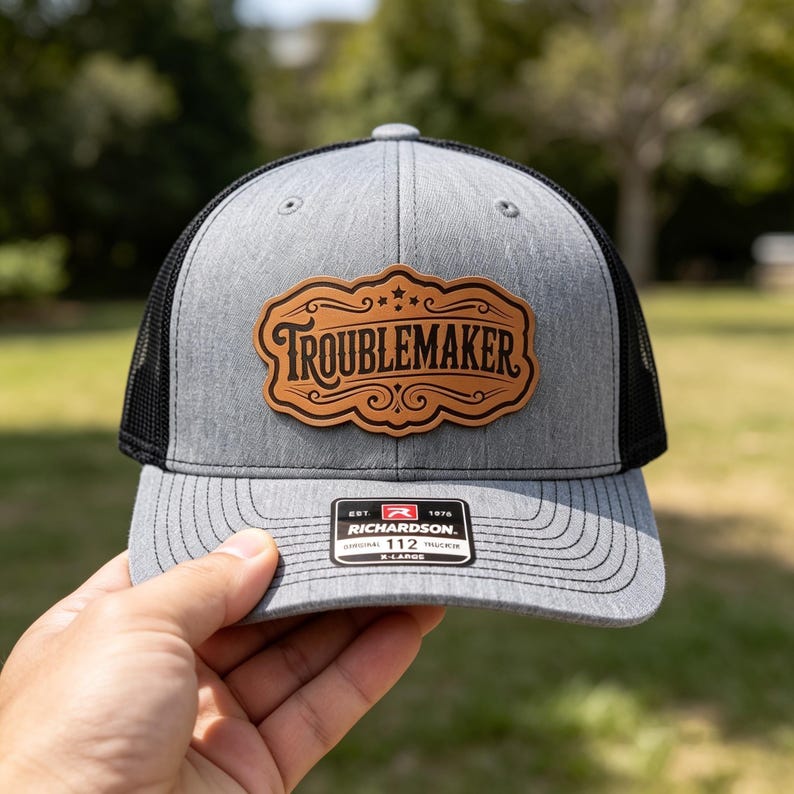 Pode incluir: Um bon&eacute; trucker cinza mesclado e preto com um patch de couro sint&eacute;tico bege na frente com a palavra TROUBLEMAKER em uma fonte decorativa. A aba tem um adesivo preto com o texto RICHARDSON 112 X-LARGE e outros pequenos detalhes.