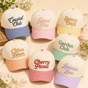 May include: Seven baseball caps with off-white crowns and gingham brims in blue, pink, yellow, orange, red, green, and purple. Each cap features embroidered cursive text: Coastal Club, Sweet Pea, Sunny Days, Citrus Bloom, Cherry Picnic, Garden Club, and Lavender Lane.