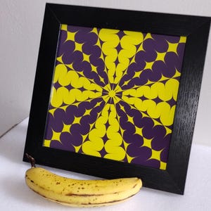 May include: A framed print with a geometric design in yellow and purple. The artwork features a radial pattern of star-like shapes. The black frame is square. A ripe banana is in the foreground.