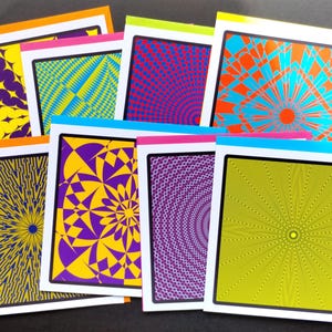 May include: A selection of greeting cards with striking geometric patterns. Each card displays a unique design in colours such as yellow, purple, blue, and orange, framed by white borders and colourful edges. The cards are stacked, presenting a variety of abstract art.
