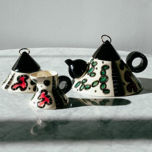 May include: A ceramic tea set consisting of a teapot, a creamer, and a lidded sugar bowl. Each piece features a cream-colored base with black conical lids, black handles, and a decorative pattern of green and red organic shapes outlined in black.