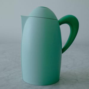 May include: A light mint green thermal carafe with a smooth, matte finish. The vessel features a rounded, dome-shaped lid, a pointed pouring spout, and a curved, slightly darker green handle attached to the side of the cylindrical body.