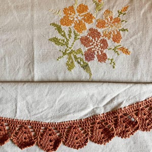 Könnte beinhalten: Ein hellbeiger Stoff mit einem Kreuzstich-Blumenmuster in Orange-, Gelb- und Grüntönen. Die Unterkante des Stoffes ist mit einer dekorativen, bogenförmigen Häkelborte in einem einfarbigen Rostorange versehen.