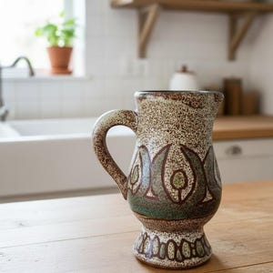 Pode incluir: Jarro de cerâmica vintage com um esmalte castanho e bege salpicado. O jarro apresenta um padrão decorativo em tons de verde e castanho. Possui uma alça e está sobre uma mesa de madeira, sugerindo utensílios de cozinha ou decoração.