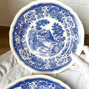 Peut inclure: Deux assiettes en céramique blanche avec un motif floral et paysager bleu. Les assiettes ont un bord festonné et un motif détaillé de fleurs et d'arbres. L'image centrale représente une scène pastorale avec des personnages près d'une rivière.