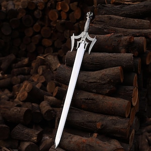 Op de afbeelding: Een zilverkleurig metalen zwaard met een lang, recht, tweesnijdend lemmet en een ingewikkelde, versierde stootplaat met puntige, vleugelachtige uitsteeksels. Het handvat is getextureerd met een gedetailleerde grip en een decoratieve pommel.