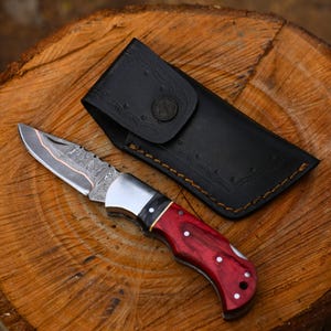 Puede incluir: Una navaja plegable con mango de madera roja y hoja estampada, junto a una funda de cuero negro. La hoja del cuchillo tiene un diseño plateado y cobrizo. La funda tiene un cierre de botón decorativo.
