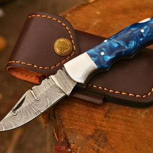 Könnte beinhalten: Ein Klappmesser mit einer Damaststahlklinge mit markantem Wellenmuster, einer Metallbacke und einem Griff aus blau marmoriertem Harz. Das Messer wird von einer braunen Lederscheide mit orangefarbenen Nähten und einem goldenen Knopf begleitet.