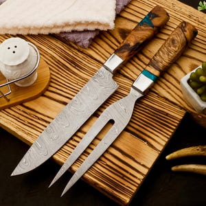 Könnte beinhalten: Ein Tranchier-Messer- und Gabel-Set mit gemusterten Stahlklingen und Holzgriffen. Die Griffe haben eine natürliche Holzmaserung mit silbernen Zwingen und türkisfarbenen Akzenten. Das Set liegt auf einem Holzschneidebrett.