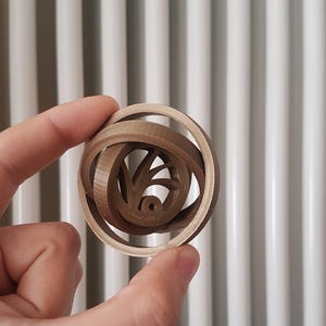 Pode incluir: Um quebra-cabeça de madeira marrom, tridimensional e interligado, segurado em uma mão. O quebra-cabeça tem uma estrutura externa circular com um design complexo no interior. O fundo é uma superfície branca com linhas verticais.