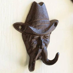 May include: A dark brown cast iron wall hook shaped like a person wearing a cowboy hat. The figure features a prominent mustache that curves upward to form two distinct hooks for hanging items. Two screw holes are visible on the hat brim.