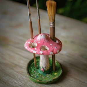 May include: A whimsical, hand-painted mushroom-shaped brush holder. The pink cap is dotted with white spots and has openings for brushes. The base is green with painted grass and a small flower. Three paint brushes are inserted into the holder.