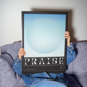 May include: A framed art print with the word "PRAISE" in white block letters on a black background. The print features a light blue and white gradient design. A small cross is below the word "PRAISE".