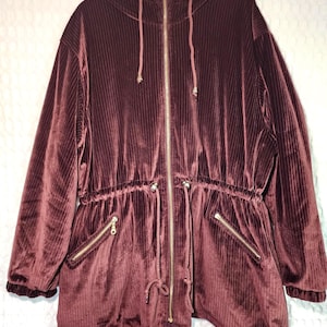 May include: A burgundy corduroy jacket with a hood and a full-length gold zipper. The jacket has two front pockets with gold zippers and a drawstring waist. The jacket's corduroy fabric has vertical ridges.