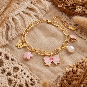 Könnte beinhalten: Vergoldetes Charm-Armband mit Schmetterling-, Blumen-, Schleifen-, Herz- und Perlenanhängern. Das Armband hat ein Kettenglied-Design und wird auf einem beigen Stoff präsentiert. Die Charms sind rosa und goldfarben.