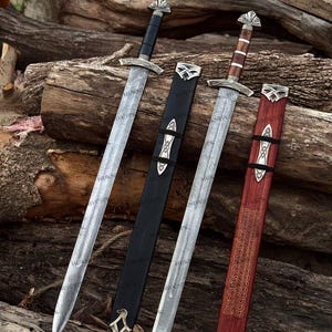 May include: Two double-edged swords with patterned steel blades and silver-toned pommels and crossguards. One sword has a black handle and black scabbard, while the other has a brown wooden handle and a red wooden scabbard with etched designs.