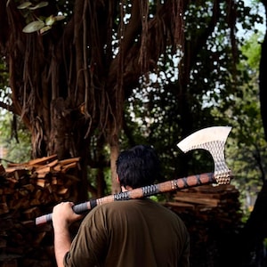 May include: A long-handled axe featuring a silver-colored metal head with a hammered, textured finish and a wooden handle wrapped with dark cord accents at the grip and near the head, held horizontally across the shoulders.