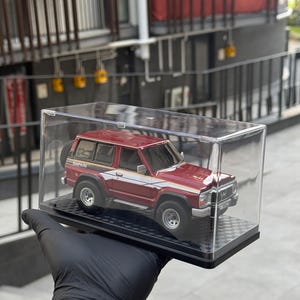 Puede incluir: Un modelo de SUV rojo y blanco, etiquetado como "Safari", se exhibe en una caja transparente. El vehículo en miniatura tiene una rueda de repuesto en la parte trasera y se encuentra sobre una base negra a cuadros. El modelo es sostenido por una mano enguantada.