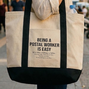 May include: A beige tote bag with black handles and a black bottom. The bag has a front pocket and text that reads "BEING A POSTAL WORKER IS EASY" with additional text below. A water bottle is in the bag's pocket.