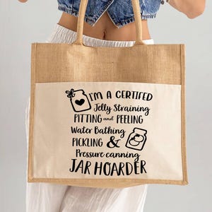 May include: A beige tote bag with a cream-colored pocket and tan handles. The bag features black text that reads "I'M A CERTIFIED Jelly Straining PITTING and PEELING Water Bathing PICKLING & Pressure canning JAR HOARDER."