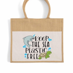 May include: A beige tote bag with a white pocket featuring a dolphin and turtle graphic. The text "Keep the Sea Plastic Free" is printed in black and blue. The bag has a natural jute texture and a brown handle.