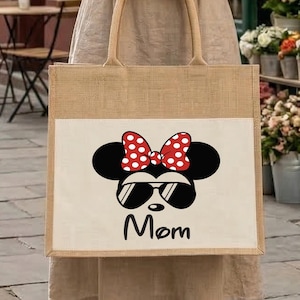 May include: A square-shaped jute tote bag featuring a white rectangular panel on the front. The panel displays a black silhouette of a mouse head wearing a red and white polka dot bow and black sunglasses, with the word Mom printed in black below.