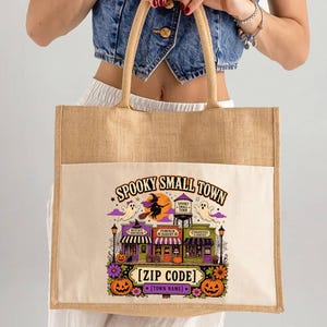 May include: A rectangular jute tote bag with a cream-colored canvas front panel featuring a colorful graphic of a spooky small town. The design includes a witch on a broom, ghosts, a water tower, and shops with the text Spooky Small Town Zip Code.