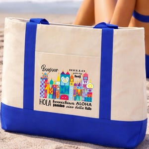 May include: A large canvas tote bag with a cream-colored body and a blue base and handles. The bag features a colorful graphic of stylized buildings and the words "Bonjour," "Hello," "Hola," and other greetings in various languages.