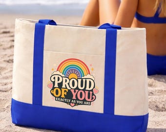Puede incluir: Un bolso tote de lona color crema con asas azul brillante y una base azul. La parte frontal presenta un gráfico colorido con un arcoíris, un sol y el texto "PROUD OF YOU EXACTLY AS YOU ARE" rodeado de pequeños corazones rosas y estrellas.