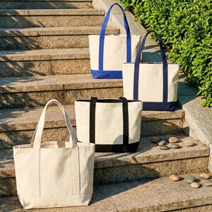 Puede incluir: Cuatro bolsas de mano de lona de diferentes tama&ntilde;os y dise&ntilde;os se muestran en escalones de piedra. Las bolsas tienen tela color crema con detalles en azul y negro. Las bolsas tienen asas resistentes.