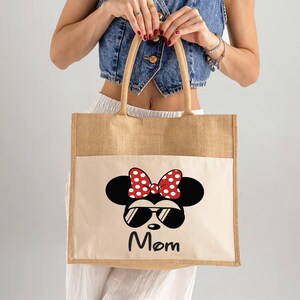 May include: A rectangular tote bag made of tan jute fabric with a cream-colored front pocket. The pocket features a graphic of a black mouse silhouette wearing red and white polka-dot bow and black sunglasses, with the word Mom printed below.