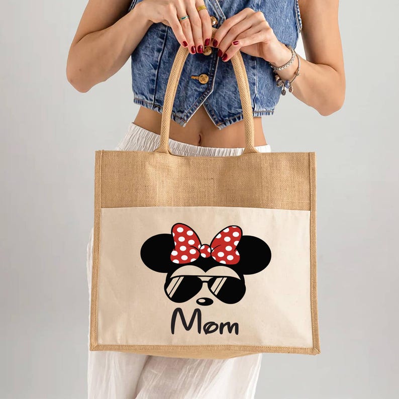 May include: A rectangular tote bag made of tan jute fabric with a cream-colored front pocket. The pocket features a graphic of a black mouse silhouette wearing red and white polka-dot bow and black sunglasses, with the word Mom printed below.