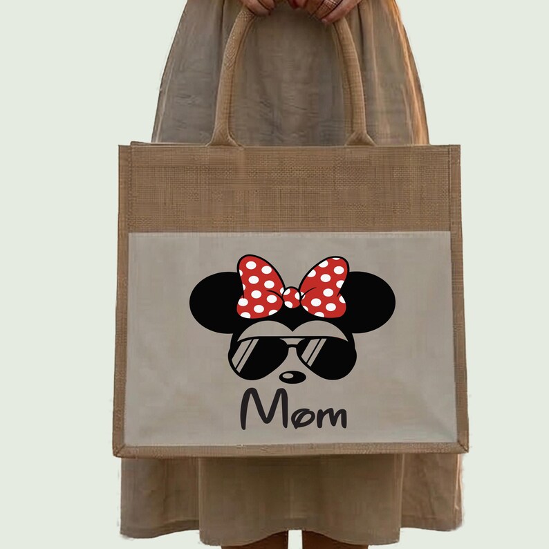 May include: A tan jute tote bag featuring a rectangular light beige front pocket. The pocket displays a black silhouette of a mouse head with a red and white polka dot bow, black sunglasses, and the word Mom printed in black script lettering.