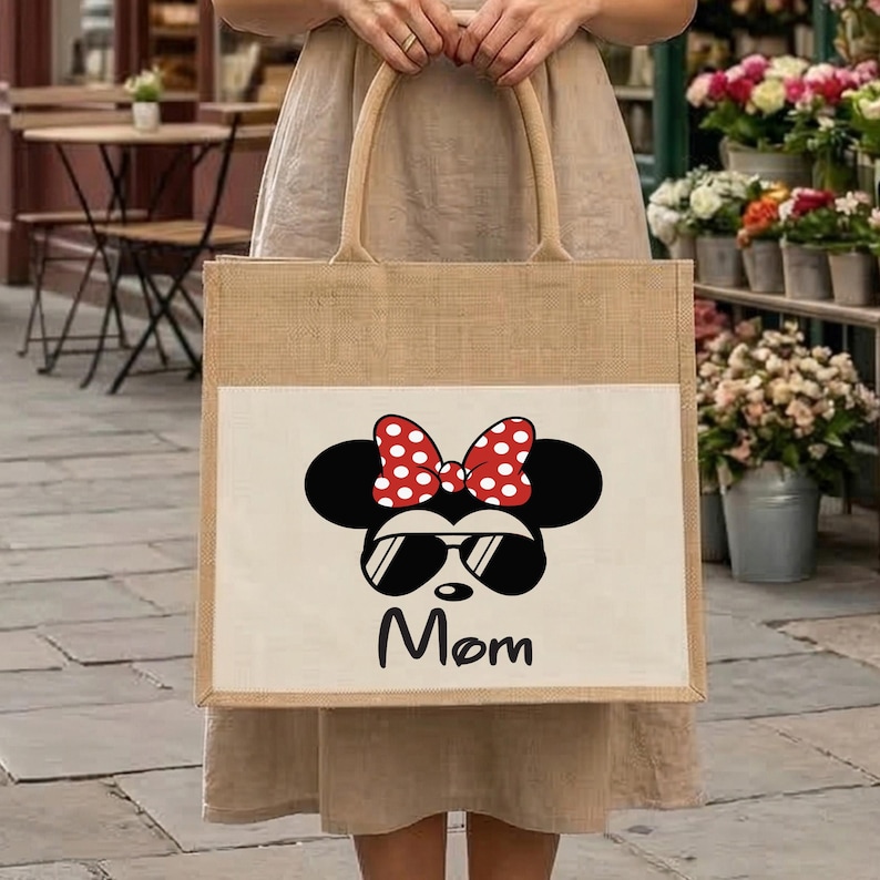 May include: A square-shaped jute tote bag featuring a white rectangular panel on the front. The panel displays a black silhouette of a mouse head wearing a red and white polka dot bow and black sunglasses, with the word Mom printed in black below.