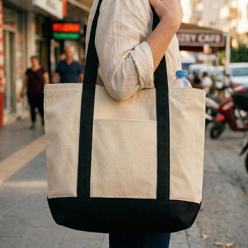 Puede incluir: Un bolso tote de lona beige con detalles en negro. El bolso tiene una base negra y correas negras. Se ve una botella de agua en el bolsillo. El bolso se lleva al aire libre.
