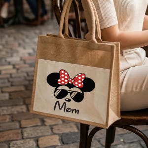 May include: A tan jute tote bag with two top handles and a rectangular white front pocket. The pocket features a graphic of a black mouse silhouette wearing red and white polka dot bow and black sunglasses, with the word Mom printed below.