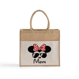 May include: A rectangular tote bag made of natural tan jute with two matching handles. The front features a white panel printed with a black silhouette of a mouse head wearing red and white polka dot bow and black sunglasses, with the word Mom below.