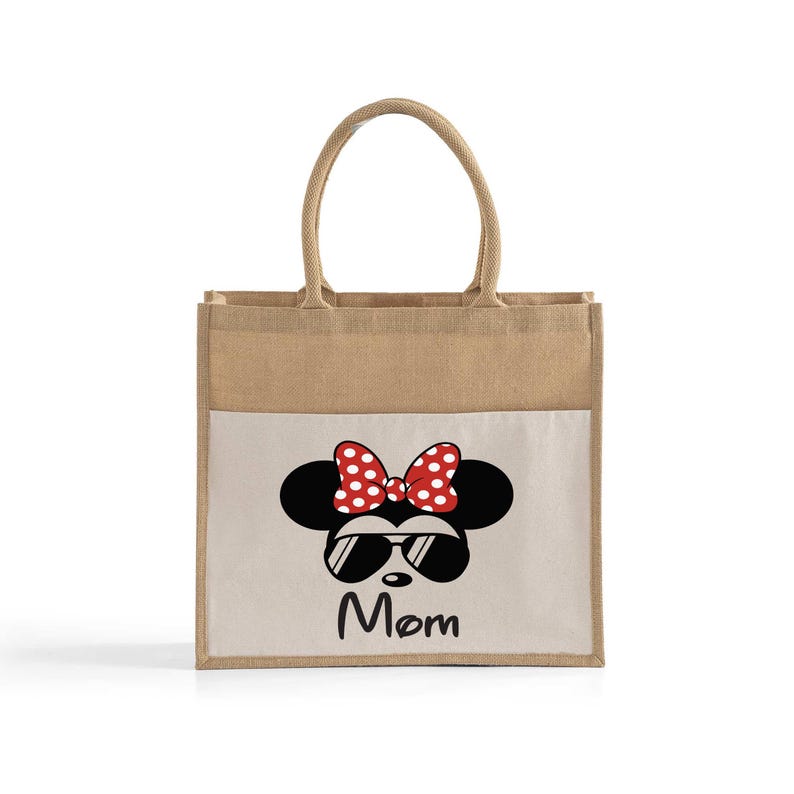 May include: A rectangular tote bag made of natural tan jute with two matching handles. The front features a white panel printed with a black silhouette of a mouse head wearing red and white polka dot bow and black sunglasses, with the word Mom below.