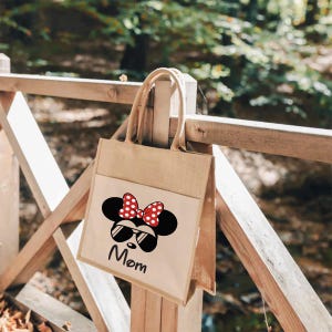 May include: A tan jute tote bag featuring a white front pocket with a black graphic of a mouse head wearing sunglasses and a red polka dot bow. The word Mom is printed in black script below the graphic. The bag has two matching tan jute handles.