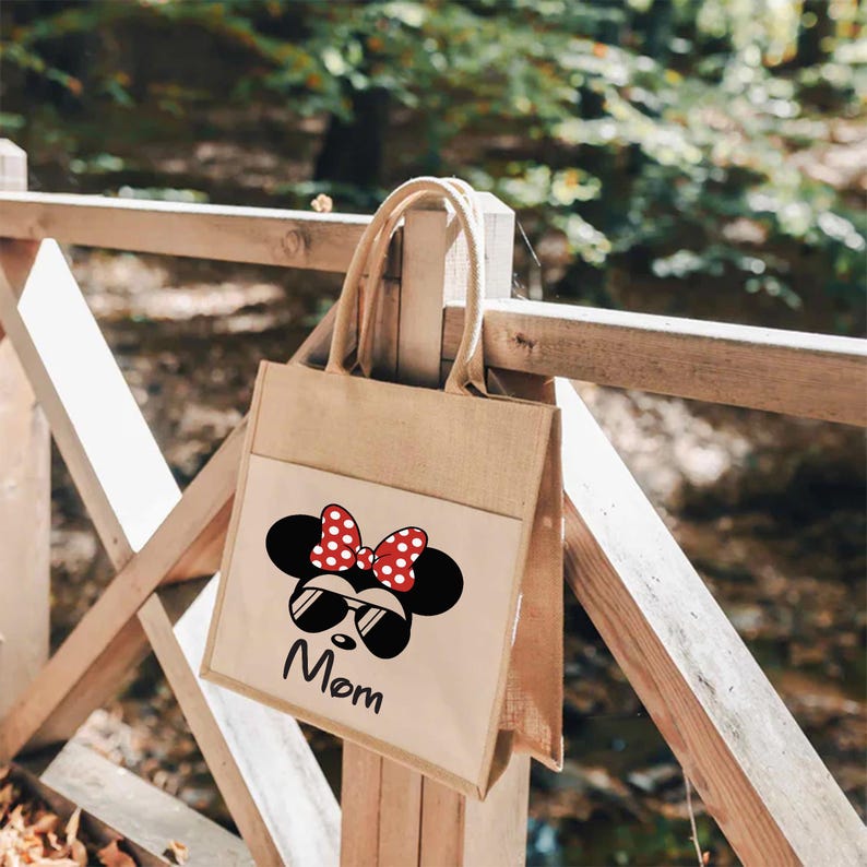 May include: A tan jute tote bag featuring a white front pocket with a black graphic of a mouse head wearing sunglasses and a red polka dot bow. The word Mom is printed in black script below the graphic. The bag has two matching tan jute handles.