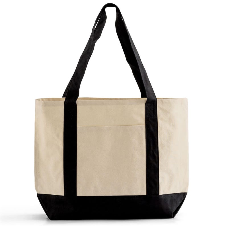 Puede incluir: Un bolso tote grande de lona con cuerpo color crema, base negra y asas negras. El bolso tiene forma rectangular con un bolsillo en la parte delantera. Las asas son lo suficientemente largas para llevarlo al hombro.