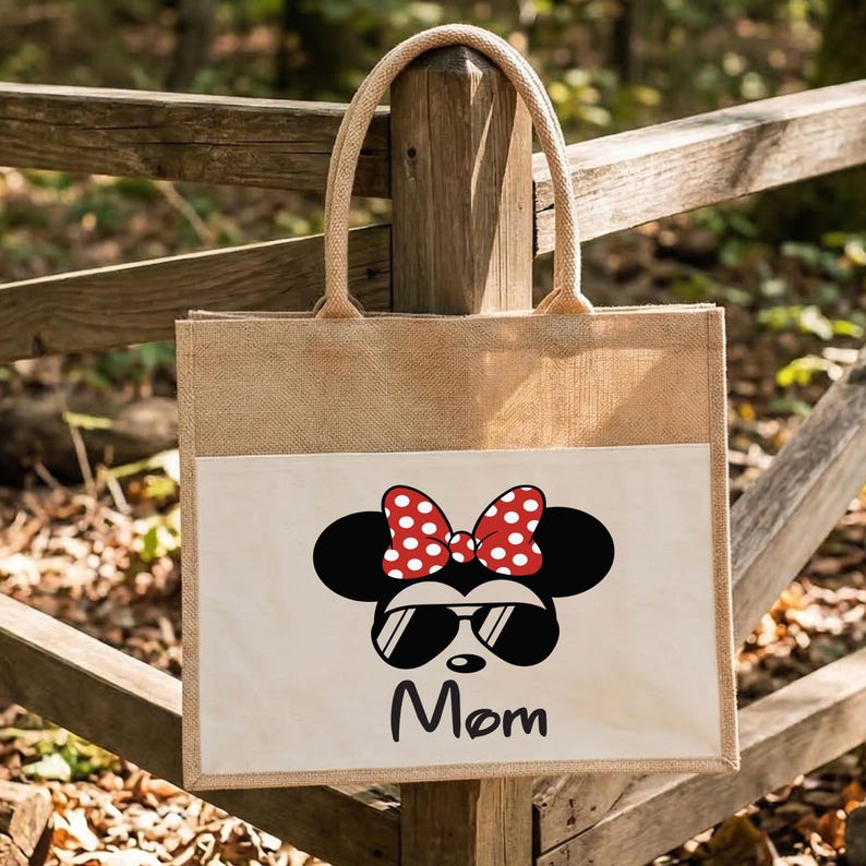 May include: A rectangular jute tote bag with two woven handles. The front features a white pocket printed with a black silhouette of a mouse head wearing a red and white polka dot bow and black sunglasses, with the word Mom written below in black.
